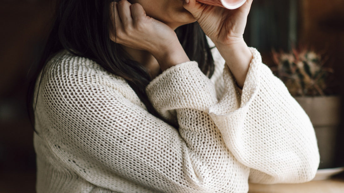 A person in a cozy sweater holding a warm mug with both hands at a wooden table
