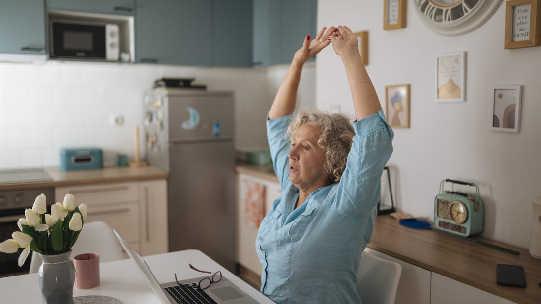 Woman sitting at her desk stretching both arms overhead in a bright home kitchen office