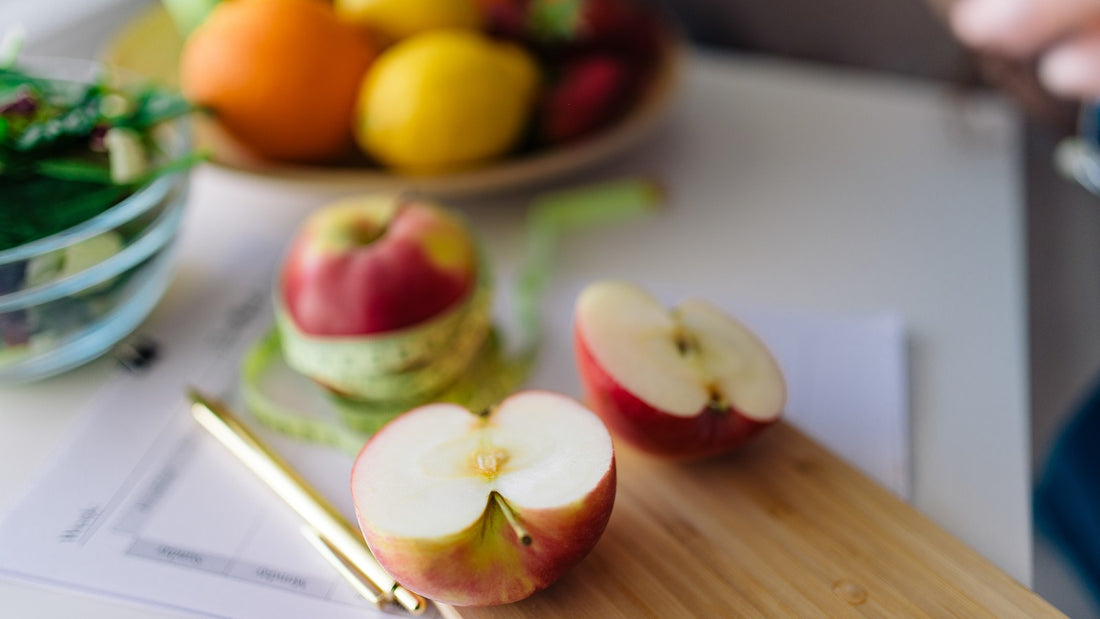 Red apple cut in half resting on a wooden cutting board next to a bowl of fresh fruit