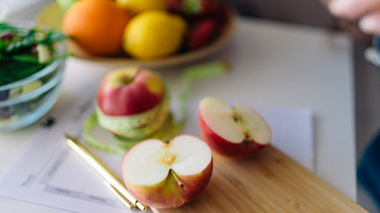 Red apple cut in half resting on a wooden cutting board next to a bowl of fresh fruit