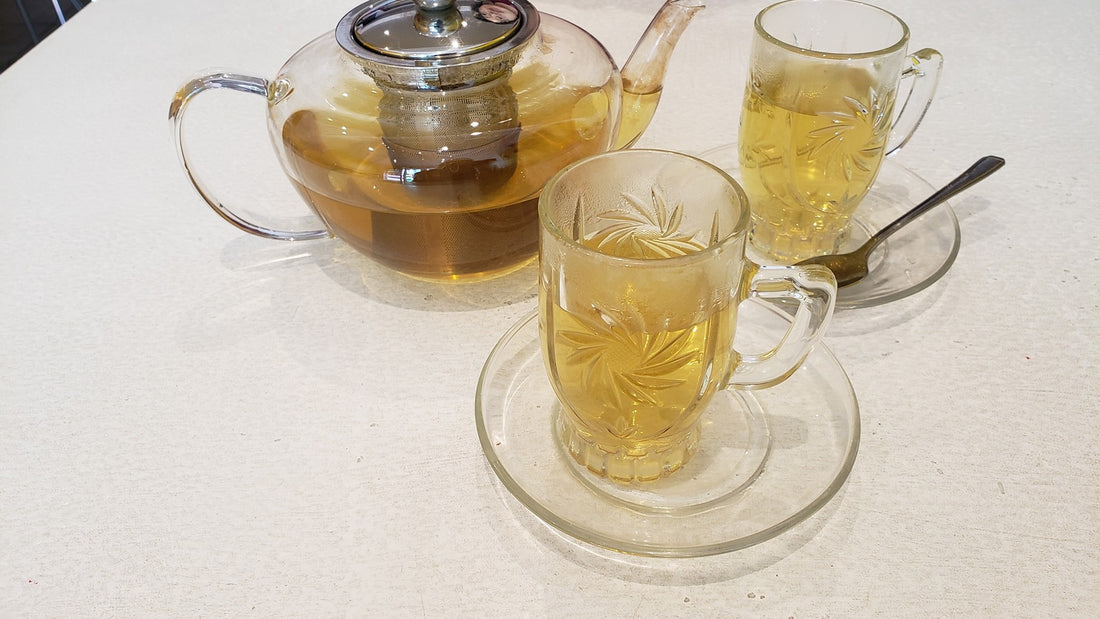 Clear glass teapot infusing golden herbal tea alongside two embossed glass teacups on a bright table