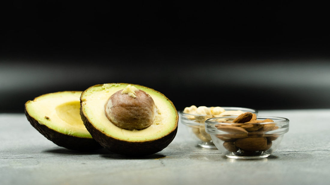 Two halves of a fresh avocado beside small glass bowls of almonds and nuts on a gray surface