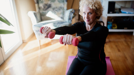 Older woman doing a seated dumbbell exercise in a bright living room