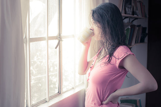 Woman in a pink shirt drinking from a mug beside a bright morning window