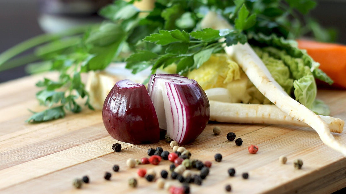 Red onion, leafy greens, root vegetables, and peppercorns on a wooden cutting board