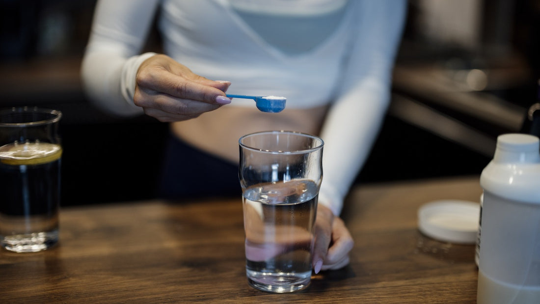 Person holding a blue measuring scoop over a clear glass of water beside an unbranded white container