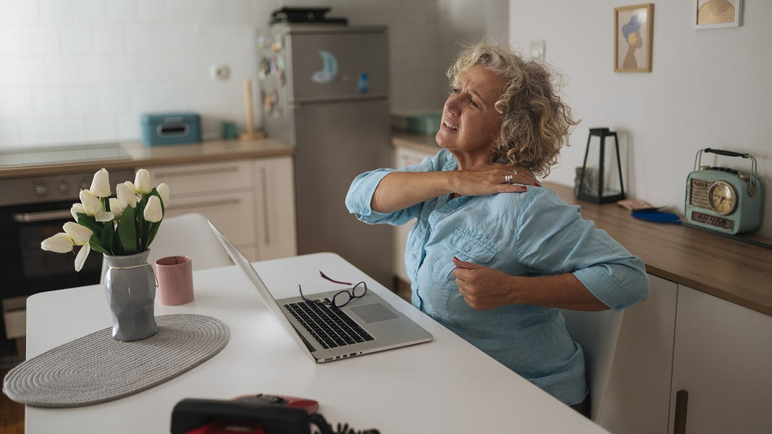 Older woman taking a seated shoulder stretch beside an open laptop