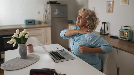 Older woman taking a seated shoulder stretch beside an open laptop
