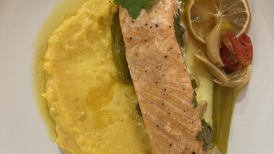 A plated grilled salmon fillet with a butternut squash puree, roasted vegetables, and lemon on a white plate
