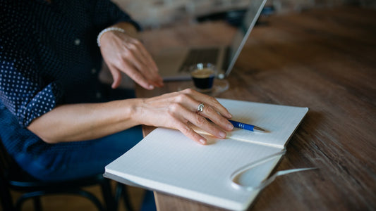Person seated at a table with an open blank notebook, pen, laptop, and coffee