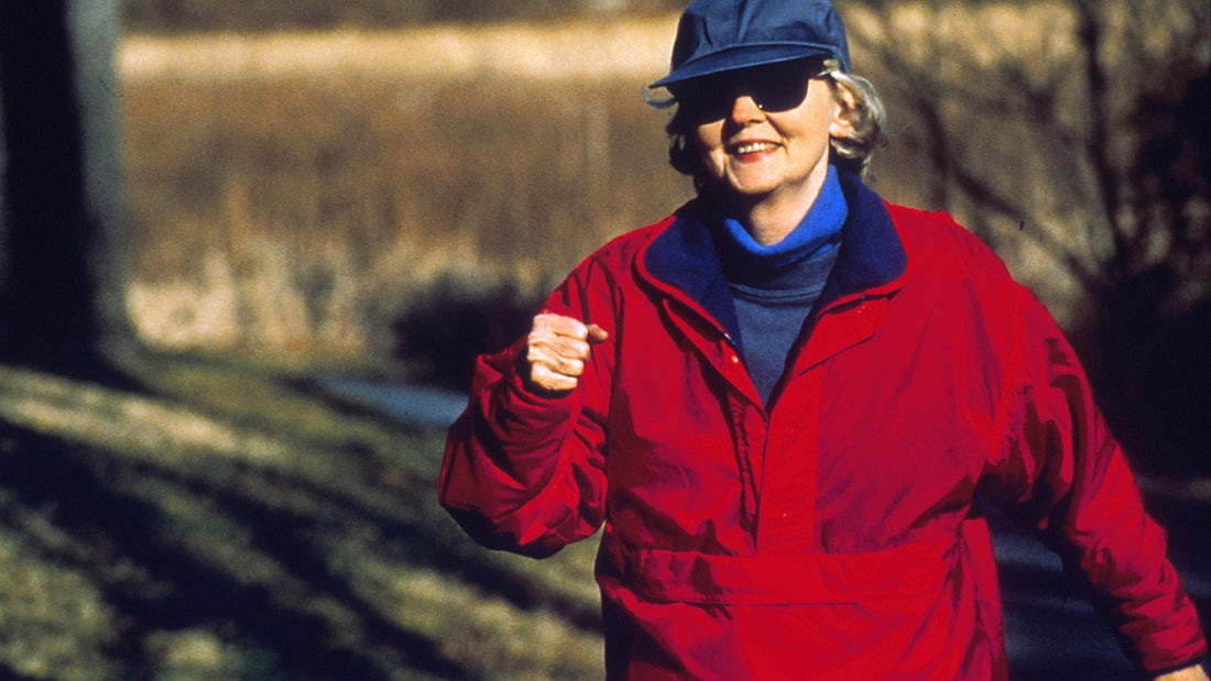 Older woman in a red jacket walking briskly outdoors on a sunny path