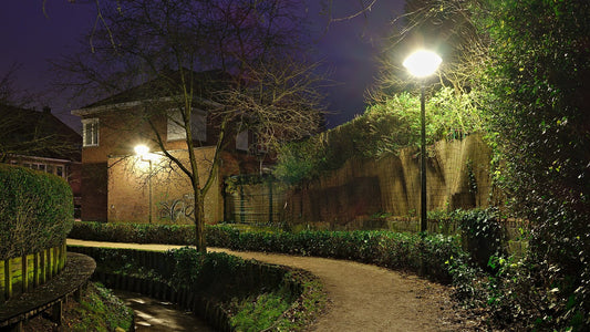 A lamplit neighborhood walking path at dusk, with trees and a bench.
